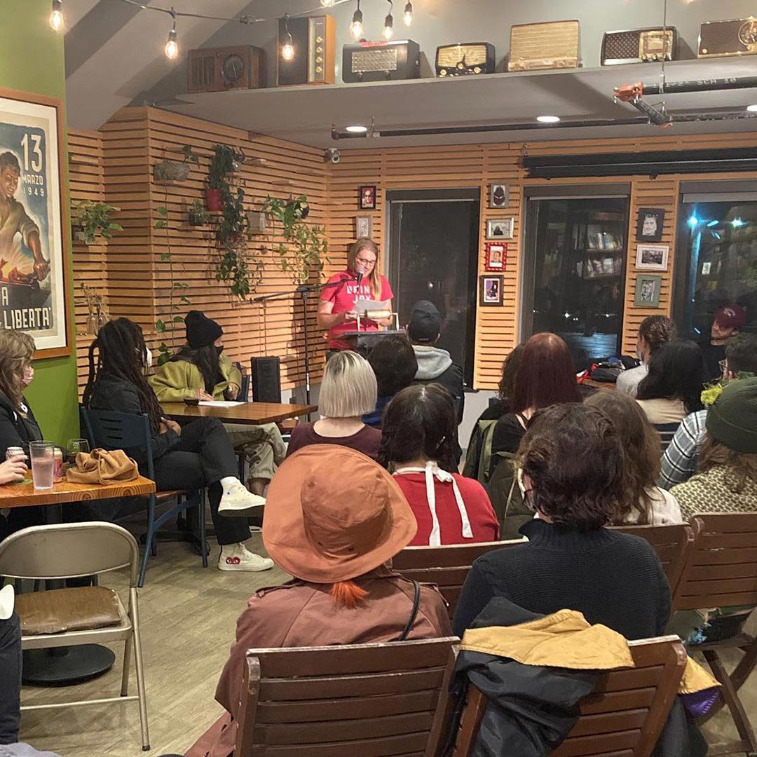 A woman is reading or speaking at a microphone in front of an audience in a cozy, warmly lit room with wooden walls. The audience is seated, listening attentively. The room decor includes vintage radios on the ceiling and various framed pictures on t