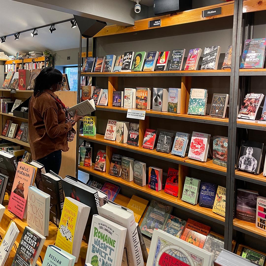 Woman browsing bookshelves in a bookstore, wearing a brown jacket and glasses, with multiple shelves of fiction and nonfiction books around her.