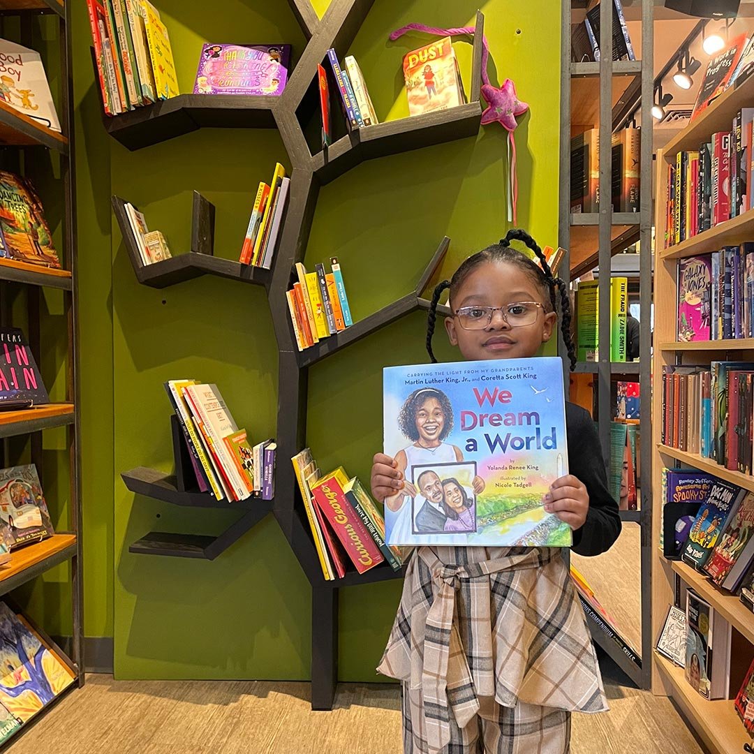 A young girl with glasses and braided hair holding a book titled 'We Dream a World' in a bookstore, with shelves of books and a green wall in the background.