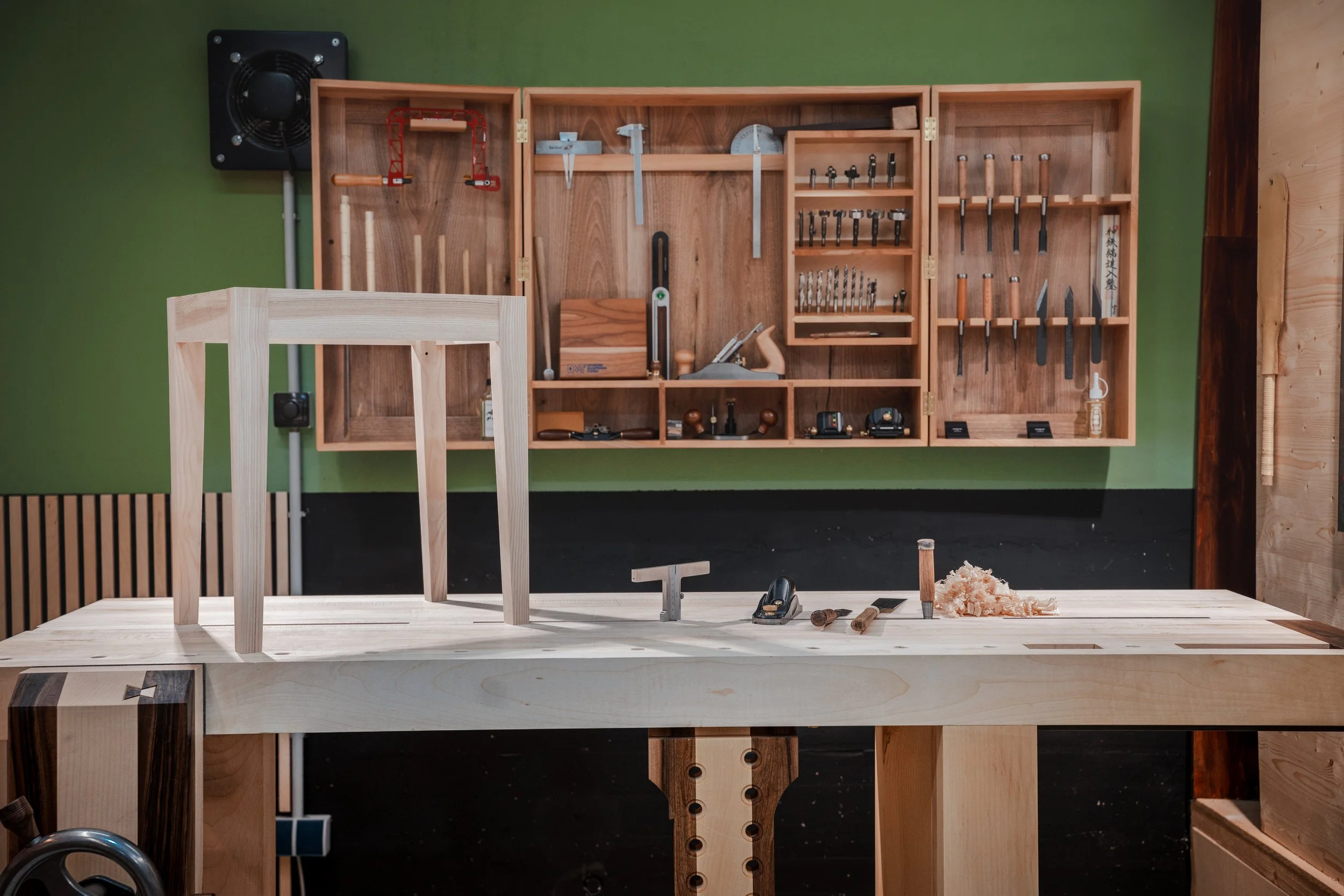 Werkbank in einer Holzwerkstatt mit verschiedenen Werkzeugen, Holzstücken und einer Holzleiste auf der Oberfläche, Wandregal mit Werkzeugen im Hintergrund.