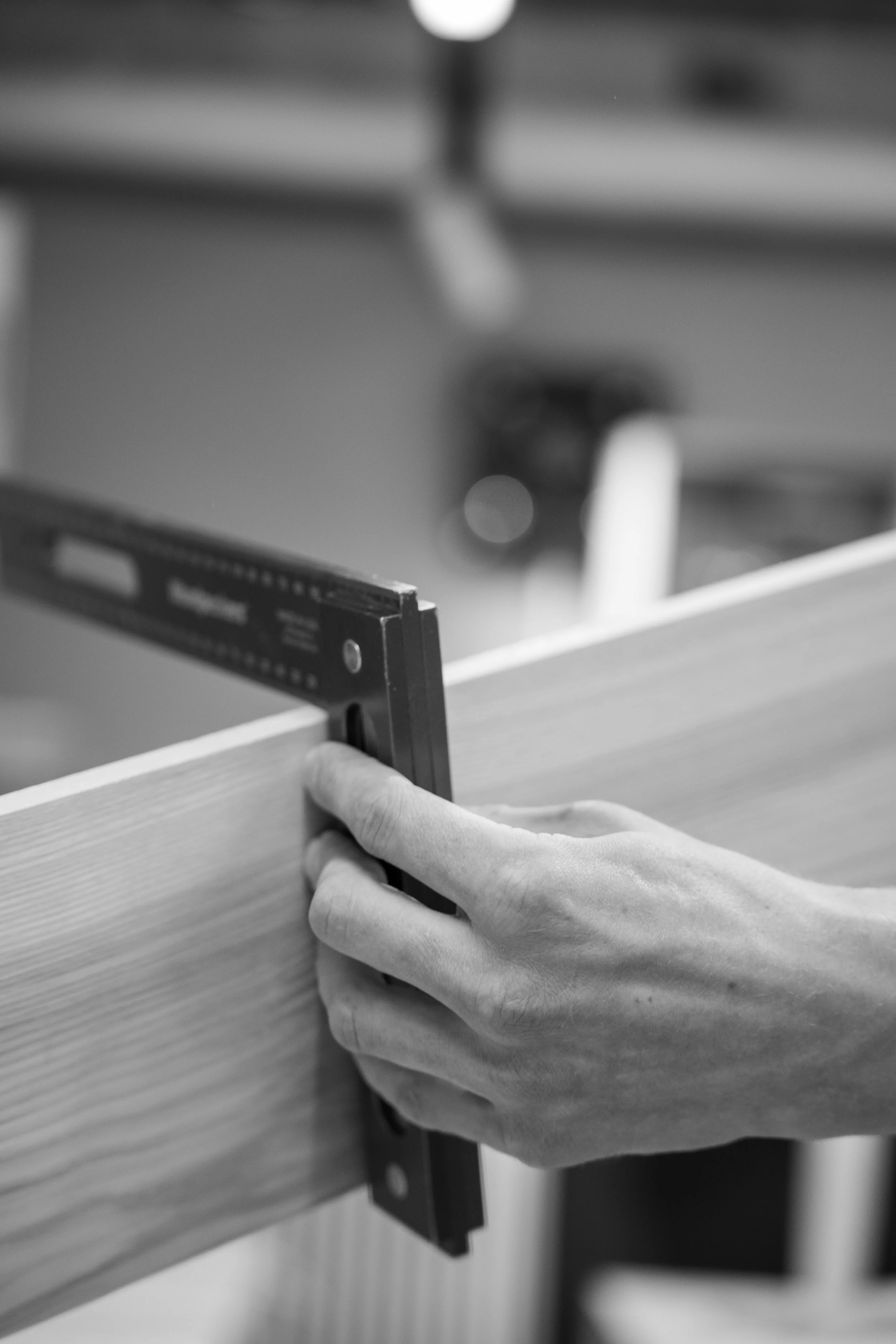 Nahaufnahme einer Hand, die eine Holzplatte mit einem Winkelmesser misst, im Hintergrund unscharf, in schwarz-weiß.