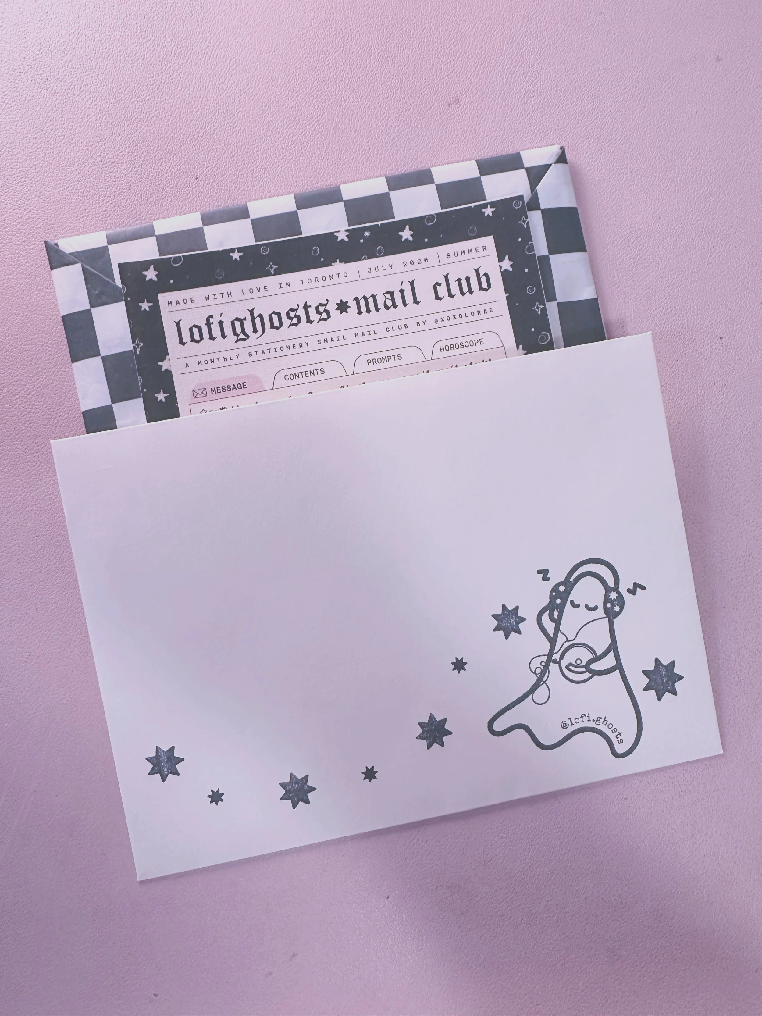 A pink surface with a white paper featuring a line drawing of a ghost holding headphones, surrounded by stars and celestial symbols.