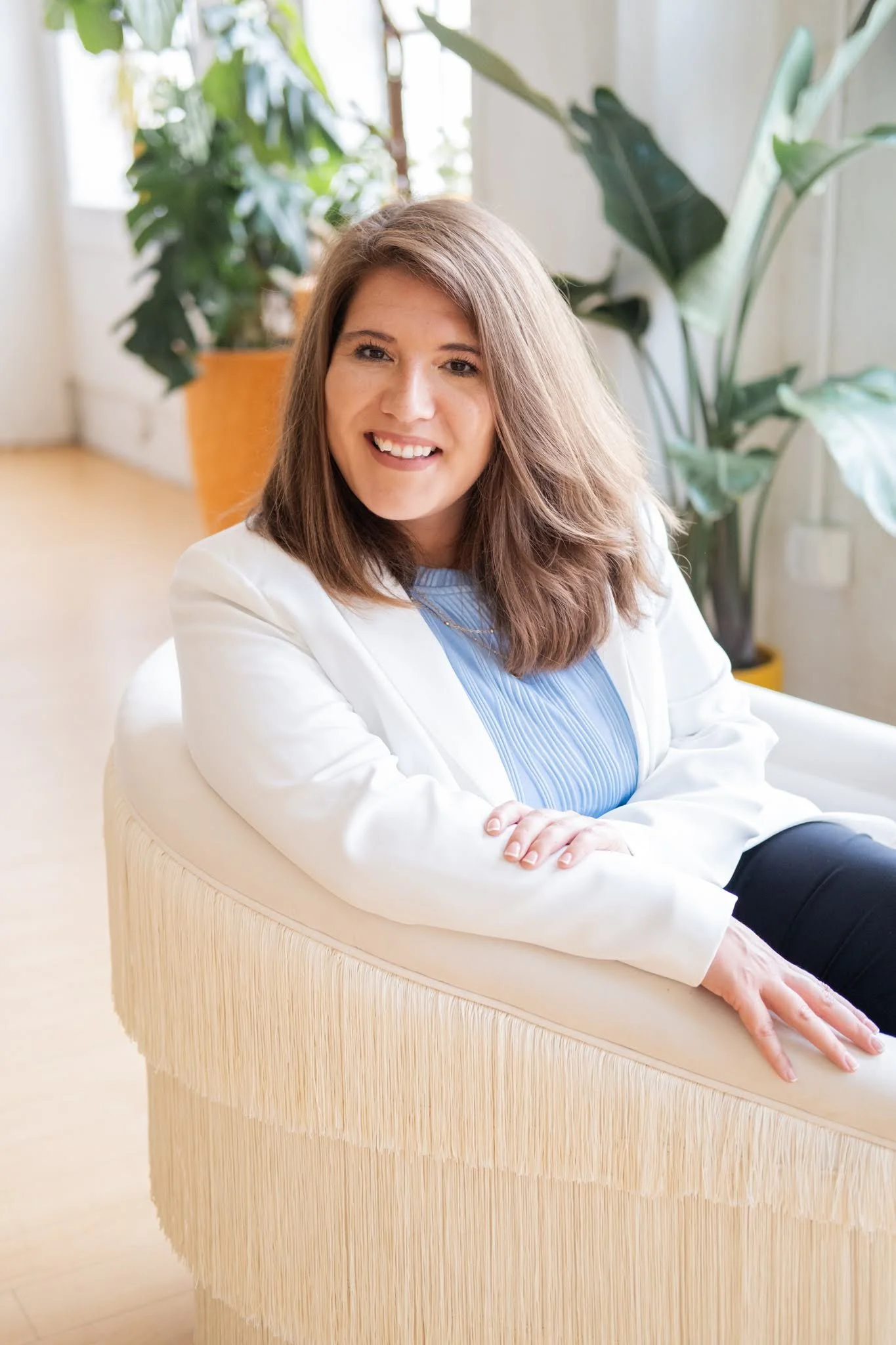 A woman with brown shoulder-length hair smiling, wearing a white blazer and a blue top, sitting on a cream-colored sofa with fringed trim, in a bright room with large green plants and natural light.