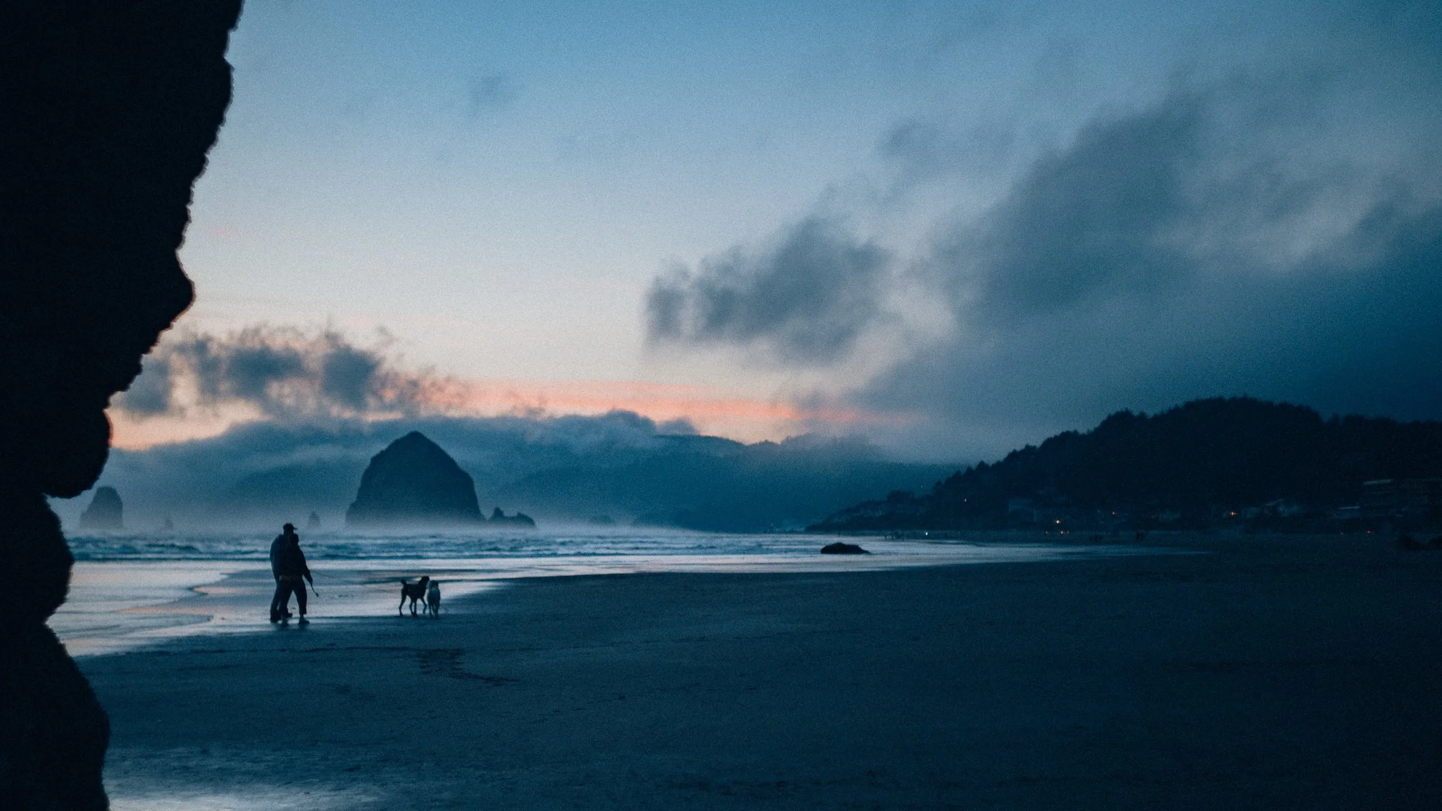 Beach at dusk with dark clouds in the sky, a person and a dog walking along the shoreline, large rock formations in the water, and distant hills.