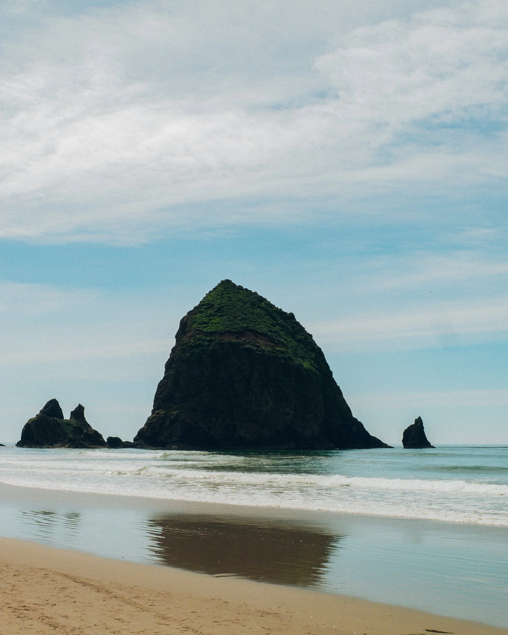 Filmic landscape photo of large volcanic sea stack with greenery, surrounded by water and smaller rocks at the coast, under a cloudy sky.