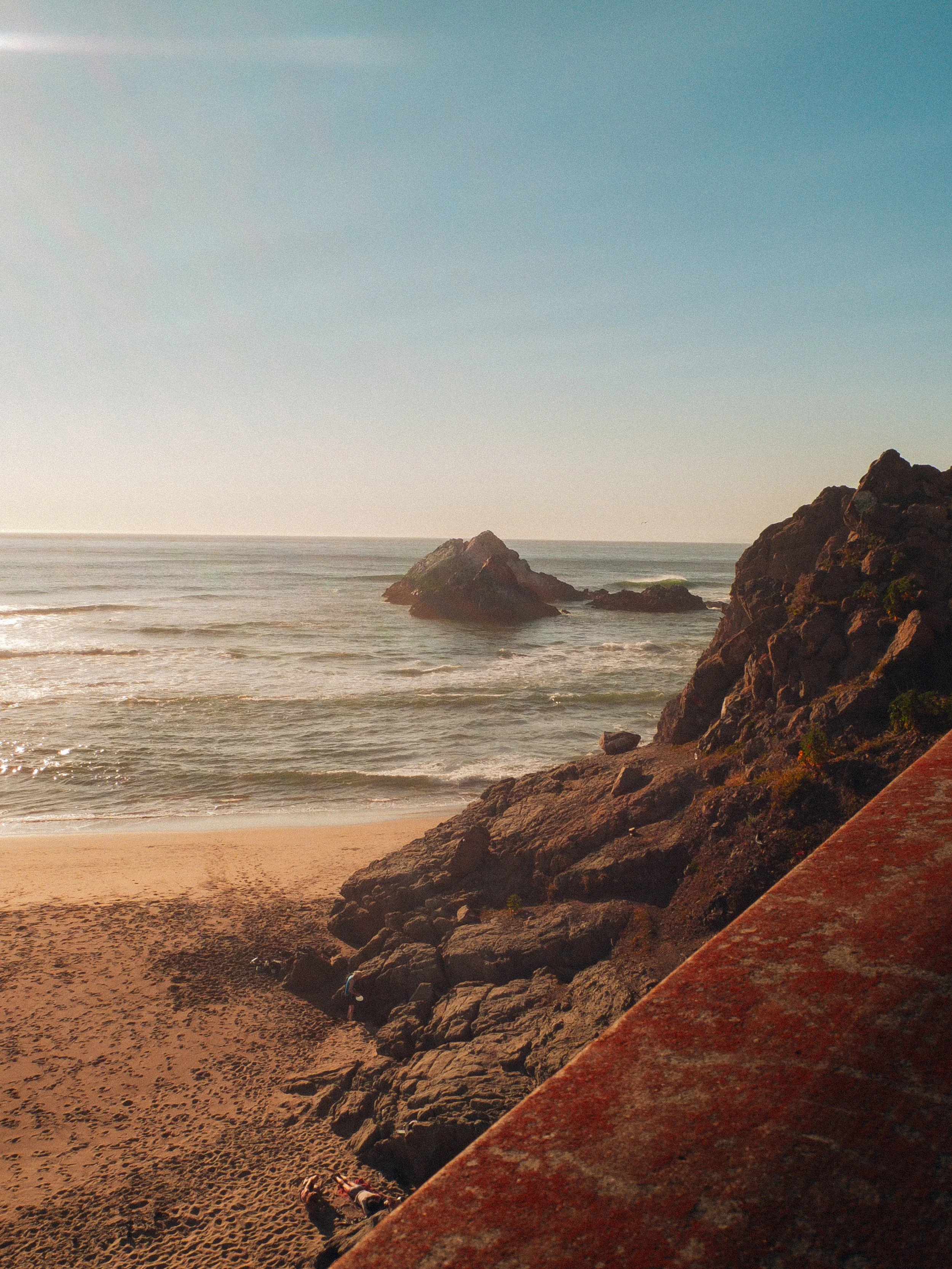 Filmic beach scene with rocky formations in the foreground, sandy beach, and ocean waves. The sky is clear with a slight gradient from light to darker blue.