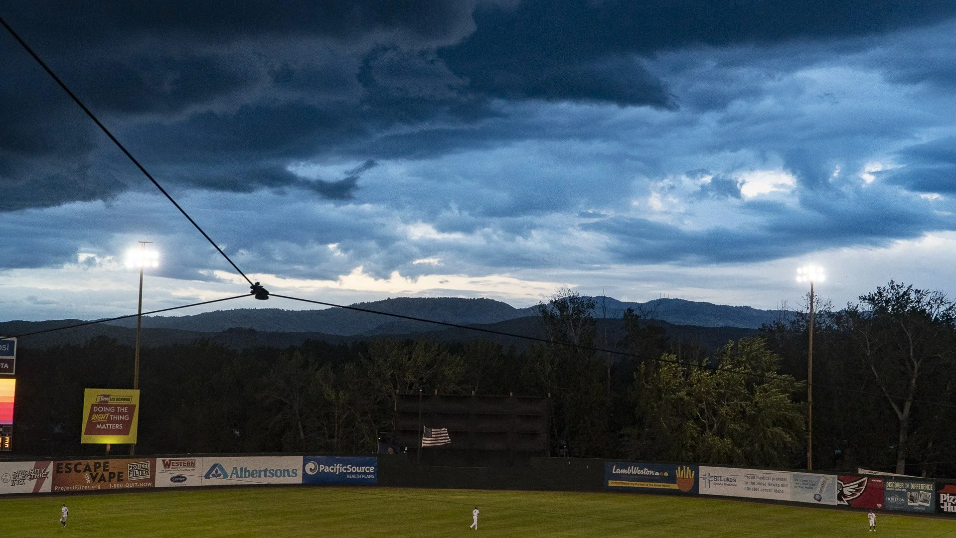 Landscape of baseball stadium field under a dark, ominous sky in evening with mountains in the background. The field has players and various advertisements along the outfield fence.