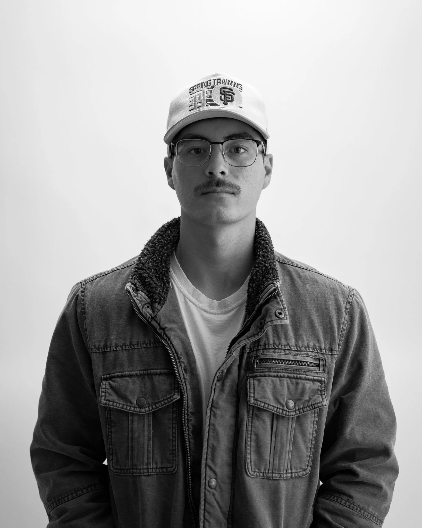 Studio portrait of young man wearing glasses, baseball cap that says "Spring Training" and collared vintage jacket against a studio backdrop.