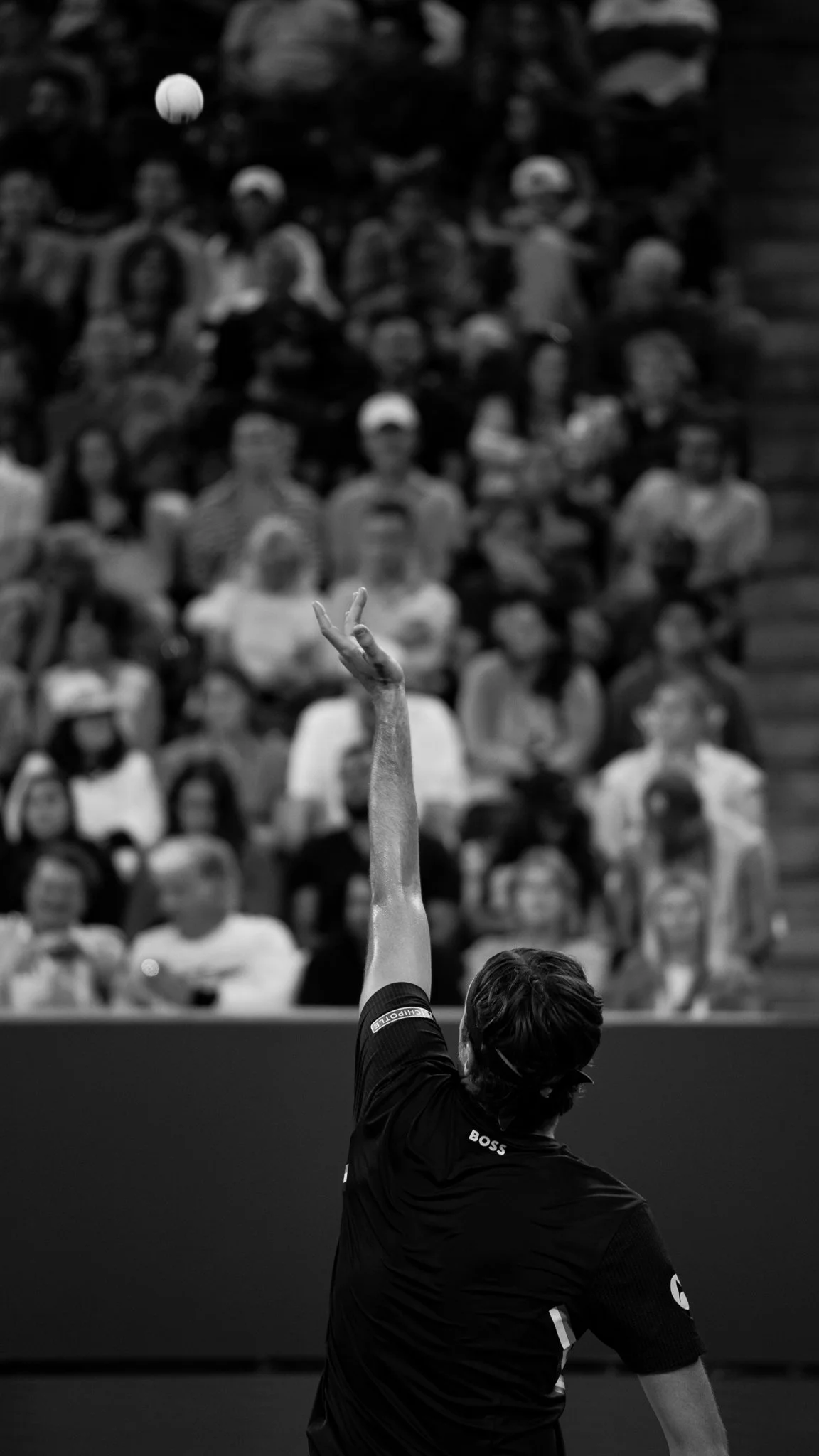 Dramatic black and white photo of tennis player tossing the ball up to serve in front of large ground.