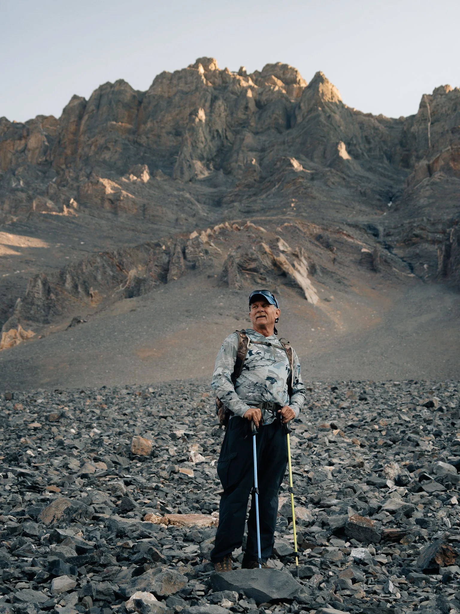 Older man in outdoor gear, holding hiking poles, standing on rocky terrain with rugged mountain in the background.