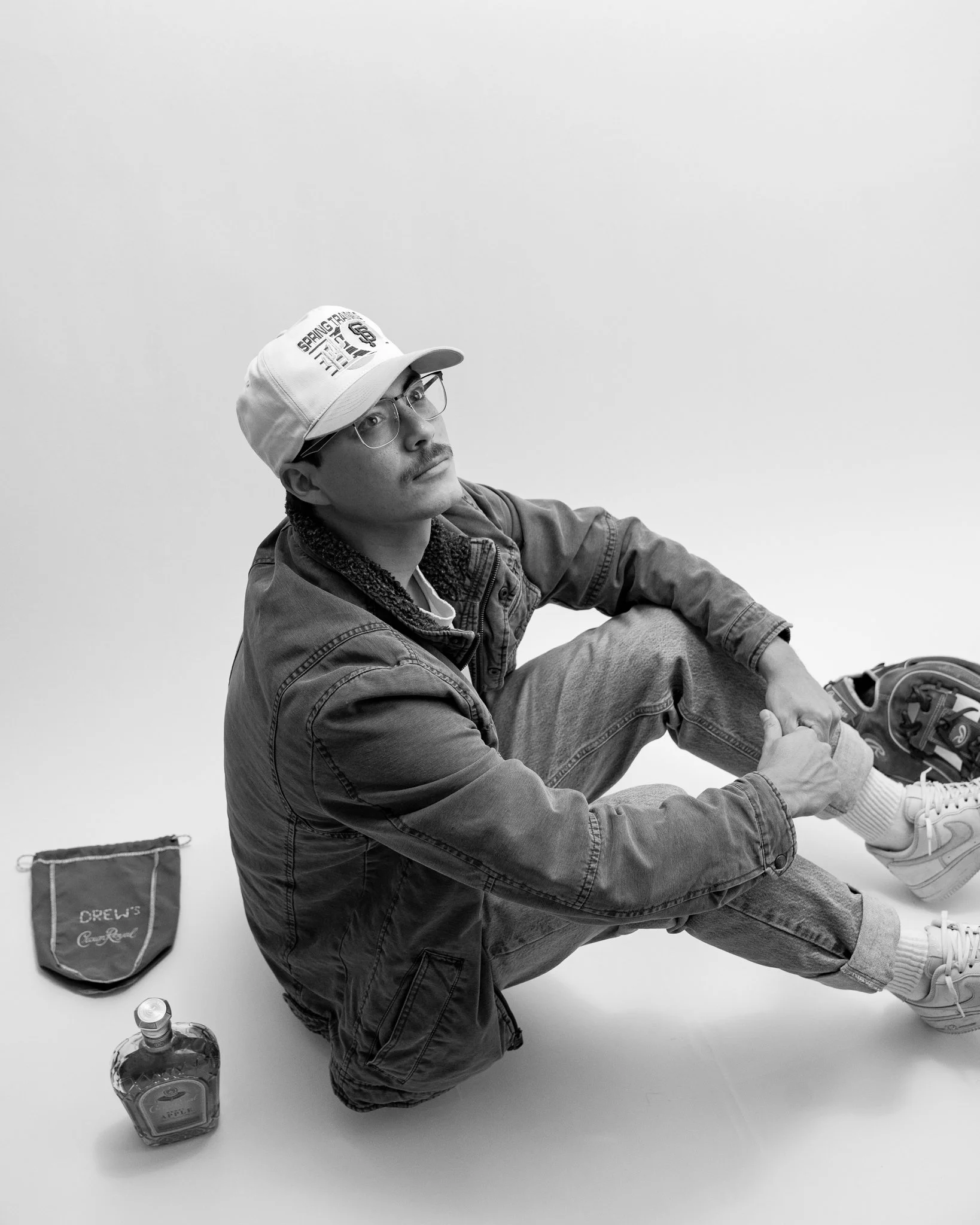 Black and white photo of a man sitting on the floor with his legs crossed, wearing glasses, a cap, and denim jacket. He looks upward with a contemplative expression. Whiskey, baseball glove, and custom bag on the floor around him. 