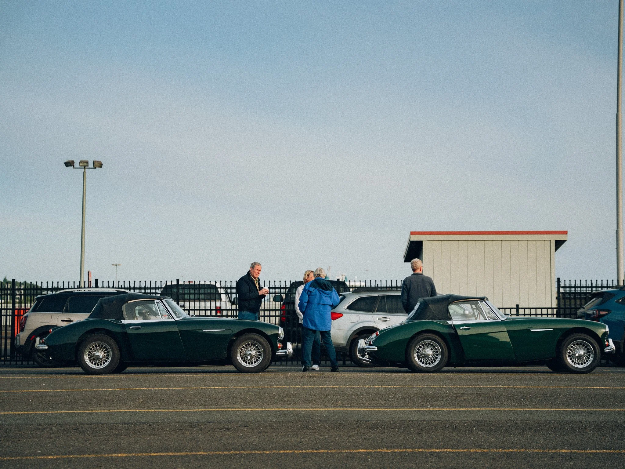 A parking lot with vintage cars and a group of people standing and talking near the vehicles. There are tall light poles and a small building with a red roof in the background, and a clear blue sky overhead.