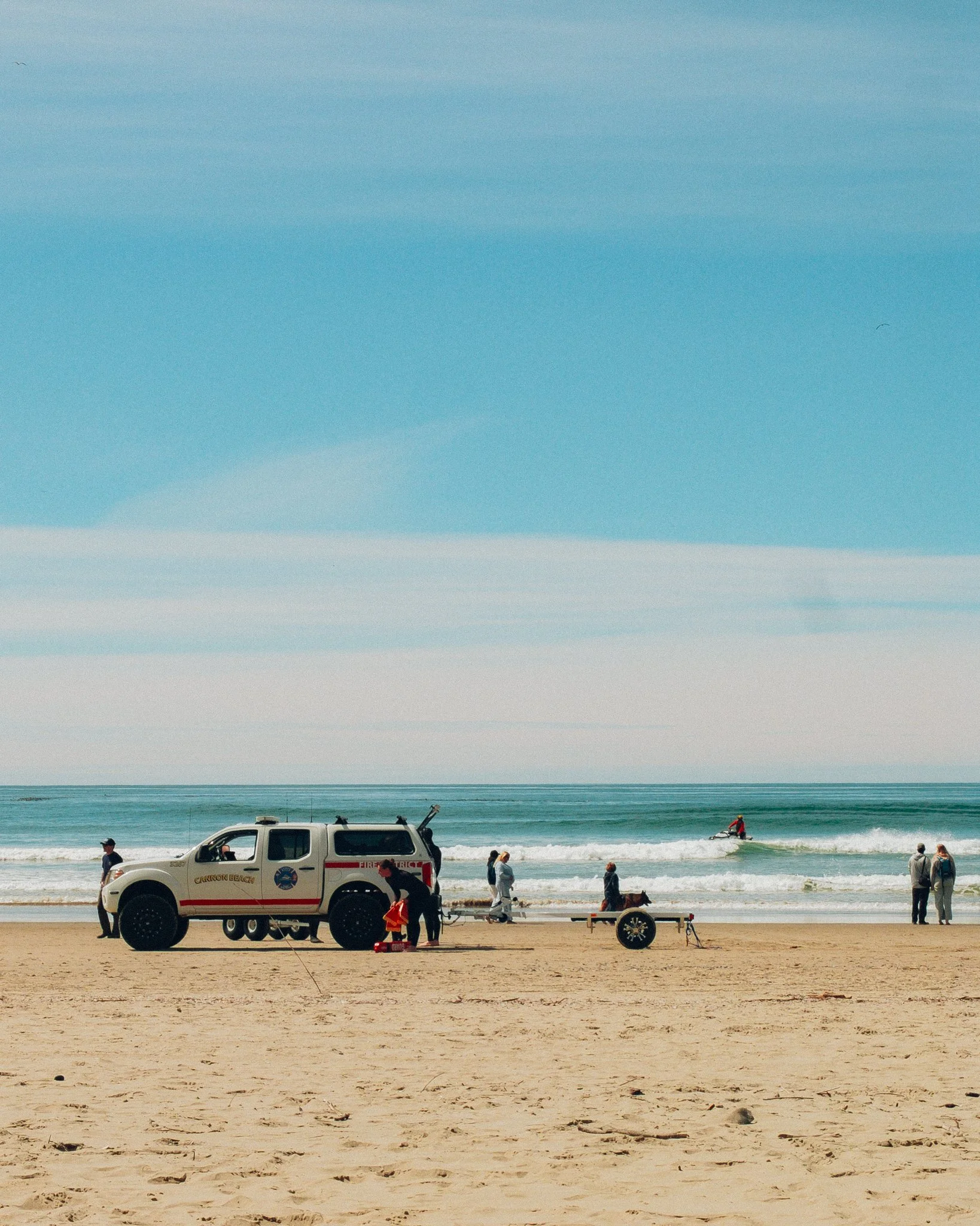 Beach scene with emergency vehicle and people along the shoreline. The sky is partly cloudy, the ocean is visible with a person riding a jet ski, and people are walking or sitting on the sand.