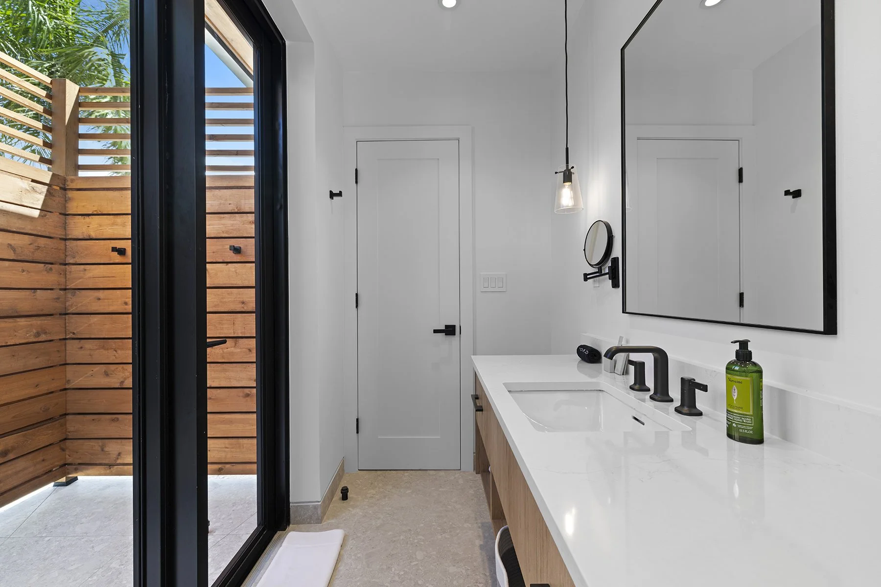 Modern bathroom with white walls, a large mirror, black fixtures, and a white countertop with a sink. An outdoor wooden privacy wall is visible through a glass door on the left, and a single pendant light hangs over the sink.