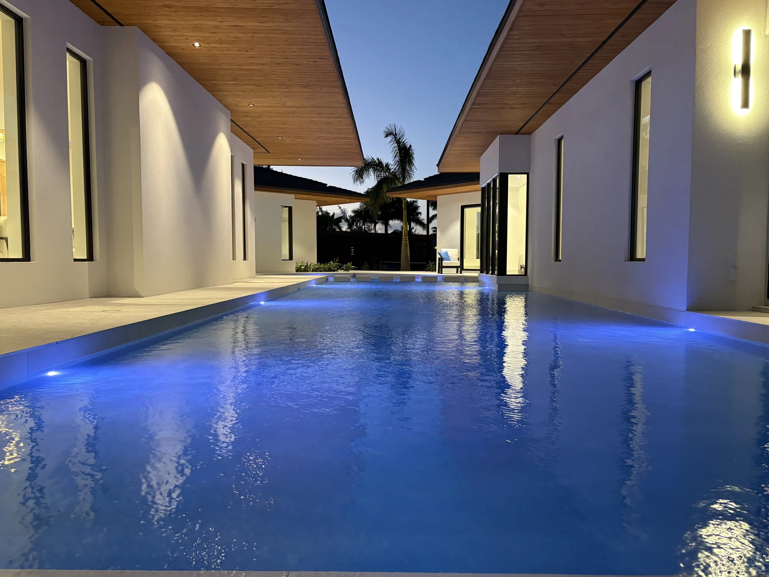 Modern house with a swimming pool illuminated by blue lights at dusk, featuring white walls, large windows, and wooden overhangs.