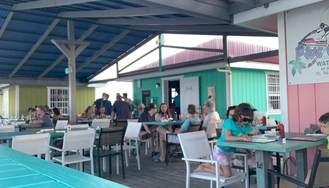 Three tacos with grilled meat, vegetables, and lemon slices on a boat dining table, with two beer bottles and condiments in the background.