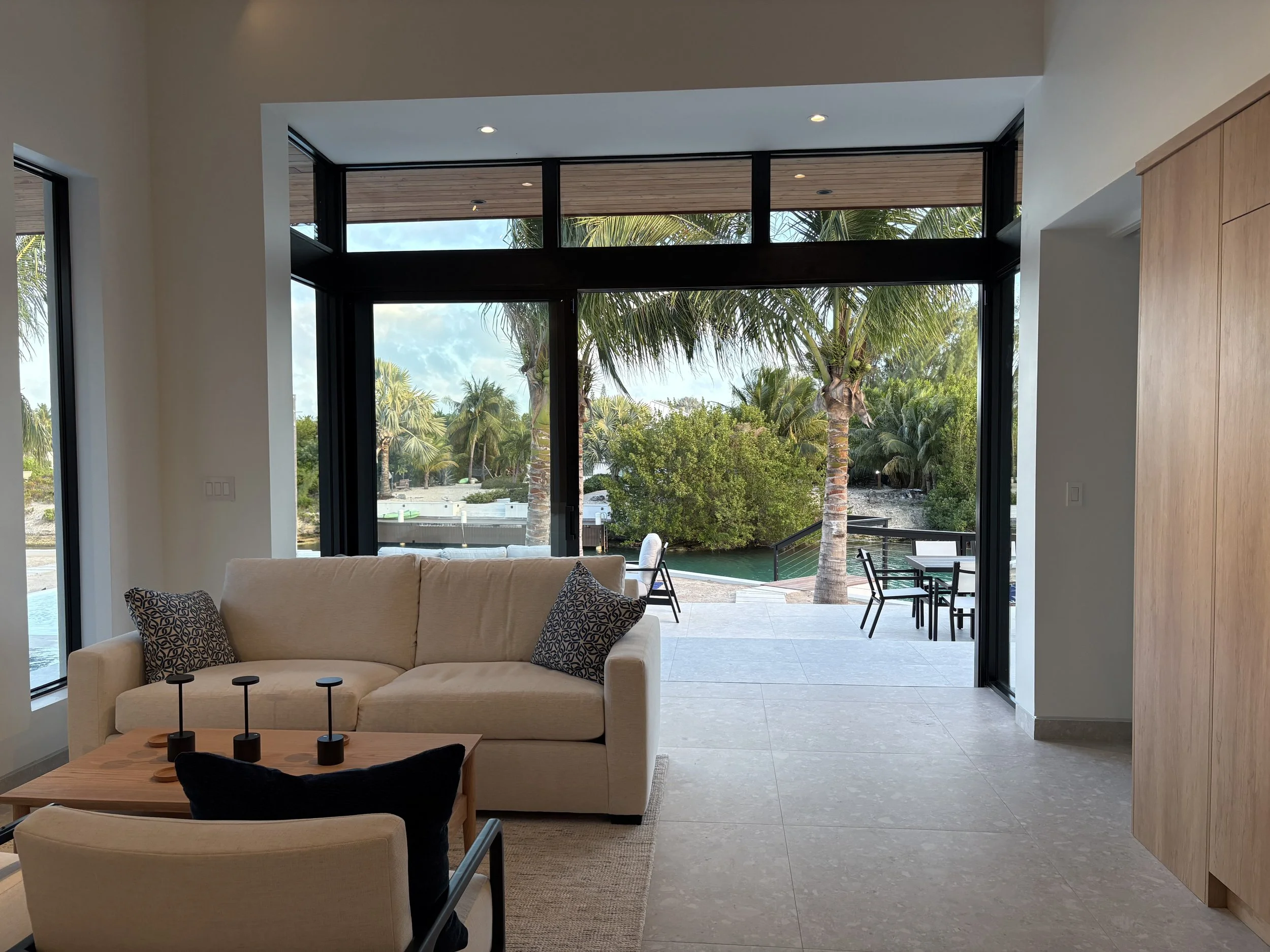 Living room with large glass doors opening to a patio with outdoor seating, overlooking a tropical landscape with palm trees and water.