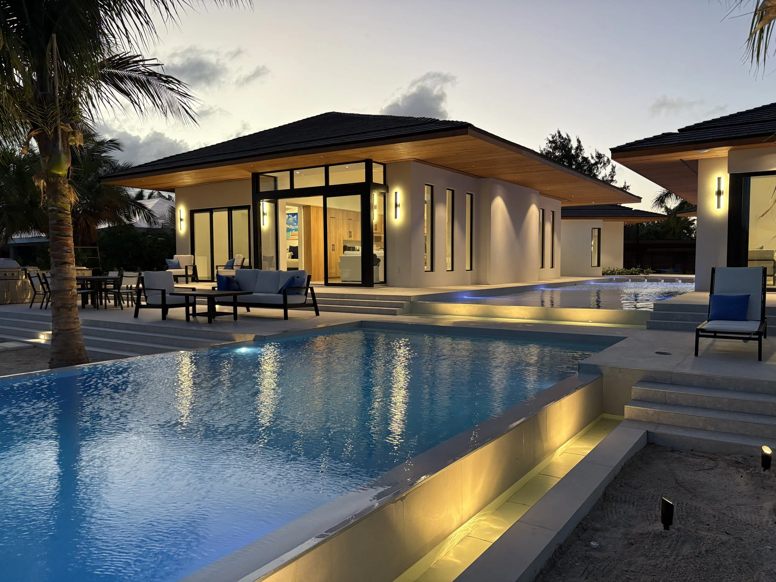Luxury modern house with illuminated windows and outdoor patio area with swimming pool, lounge chairs, and palm trees during evening.