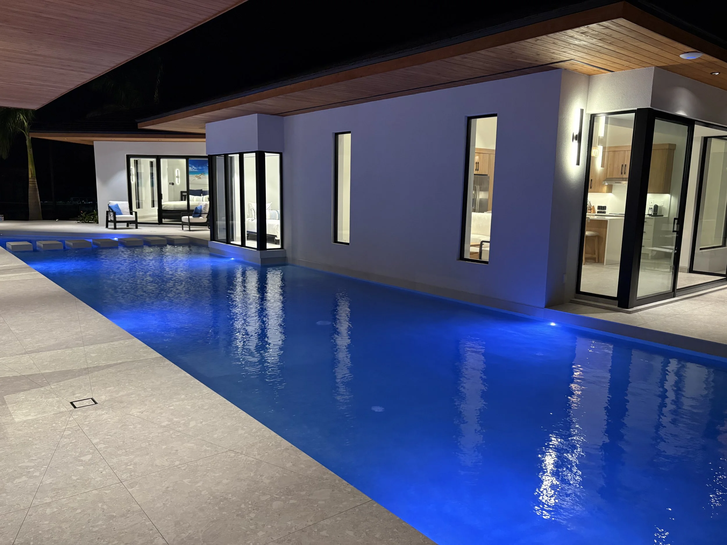 Night view of a modern house with a swimming pool illuminated by blue lights, large glass windows, and an outdoor seating area.
