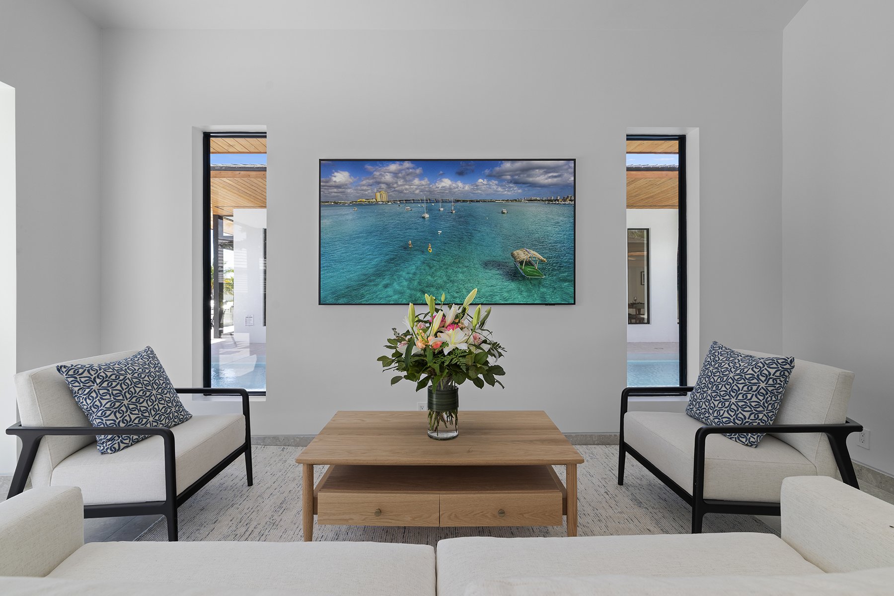 Modern living room with a wall-mounted TV, two armchairs with patterned pillows, a wooden coffee table with a flower vase, and two narrow windows showing an outdoor area with a pool.