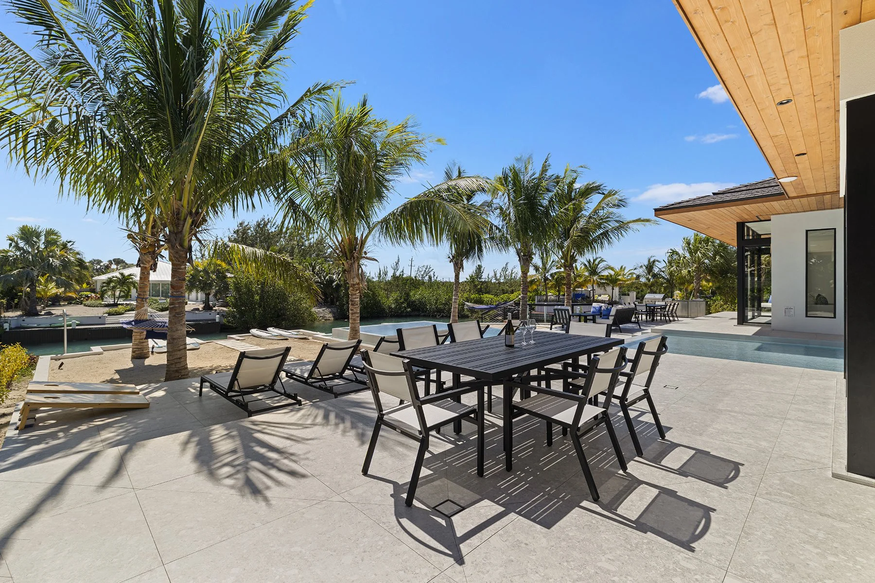 Outdoor patio with a black dining table and chairs, and lounge chairs, surrounded by palm trees, with a pool and a modern house nearby under a blue sky.