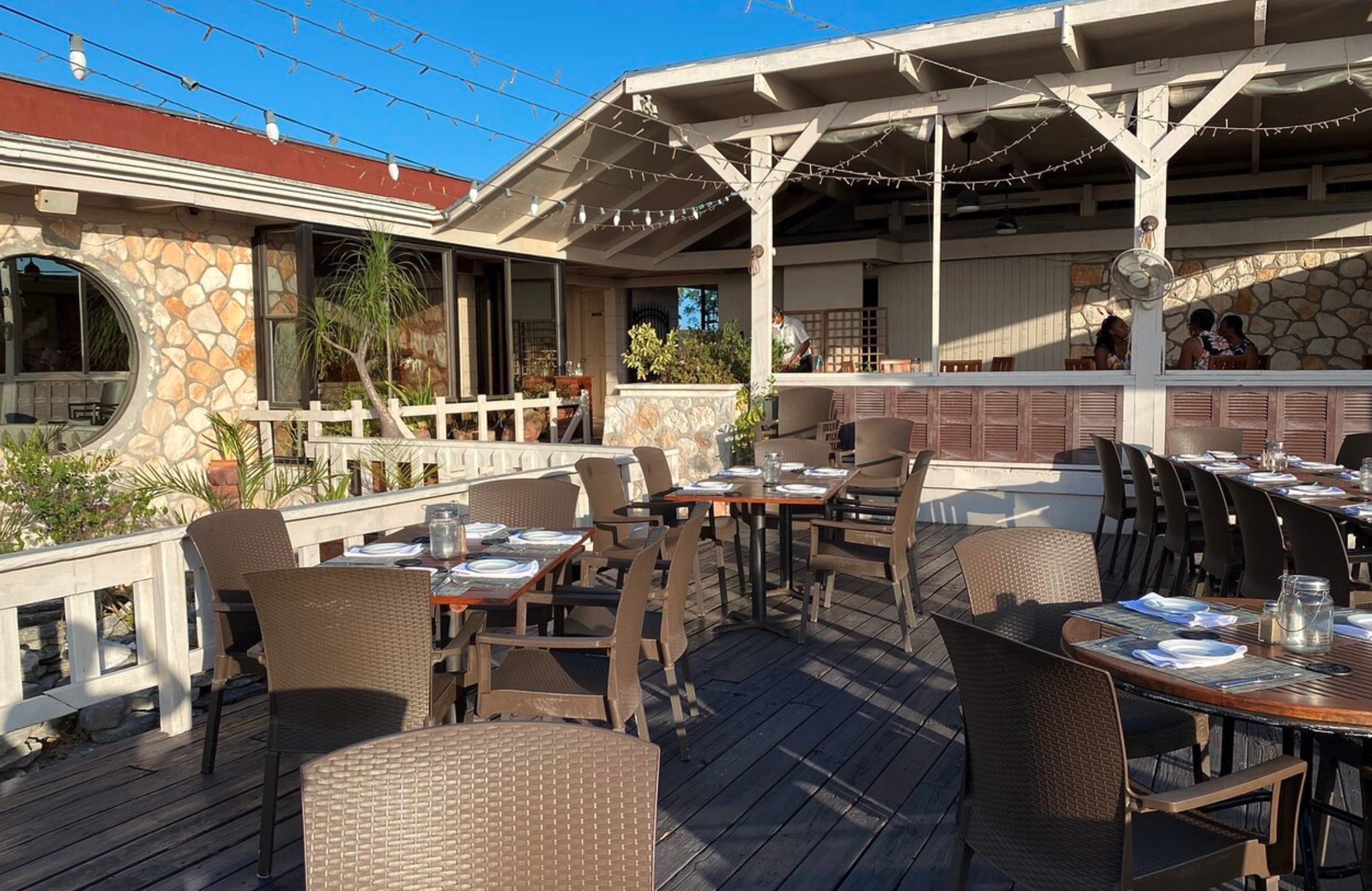 An outdoor restaurant patio with tables set for dining, featuring wooden floors, chairs, and tables with plates and glasses, surrounded by a low white fence and a stone and wood building in the background under a sunny sky.