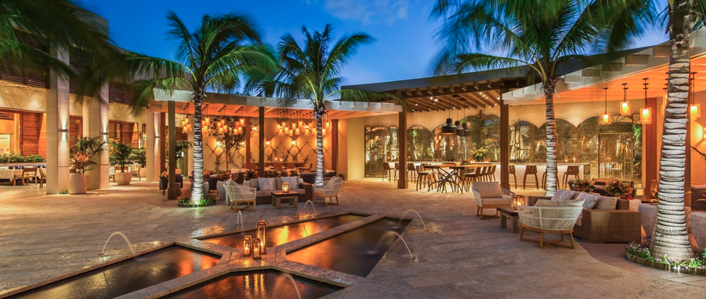 An outdoor resort with a pool area, palm trees, modern lounge furniture, and warm lighting at dusk.