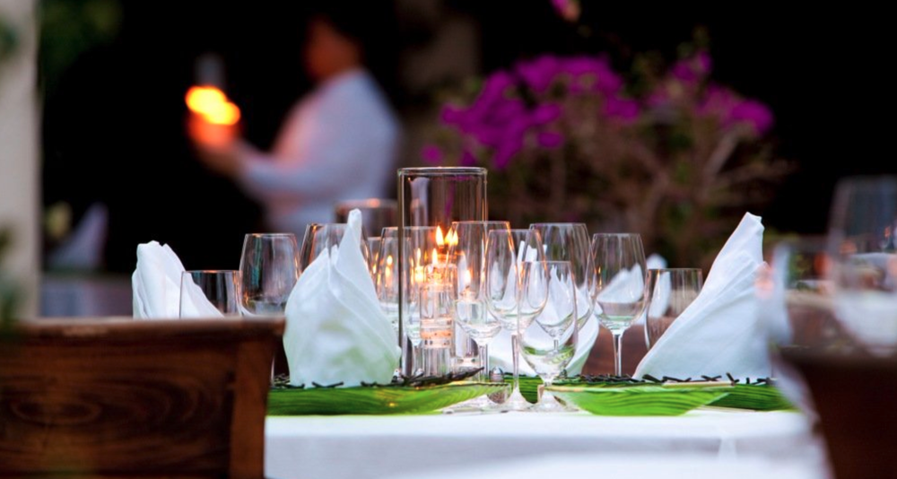 A beautifully set outdoor dining table with wine glasses, napkins, and candles, with blurred people and purple flowers in the background.