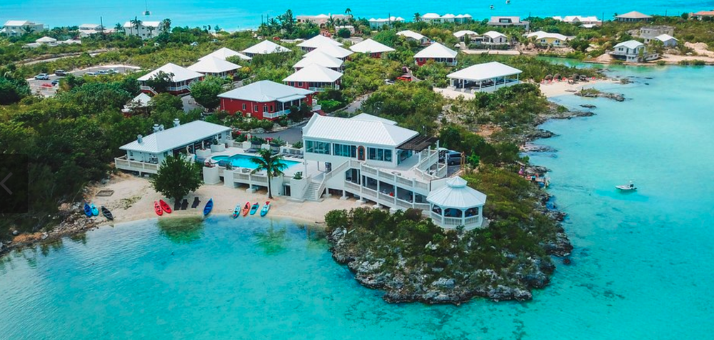 Aerial view of a coastal vacation resort with multiple houses, some with white rooftops, surrounded by lush greenery and clear blue water, featuring a small beach with kayaks and a swimming pool.
