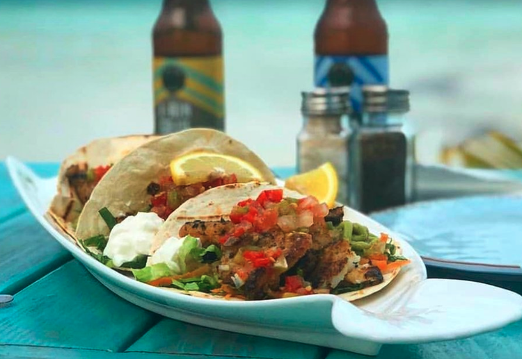 Three tacos on a white plate with lemon wedges, greens, and salsa, placed on a turquoise table with two bottles of beer in the background.