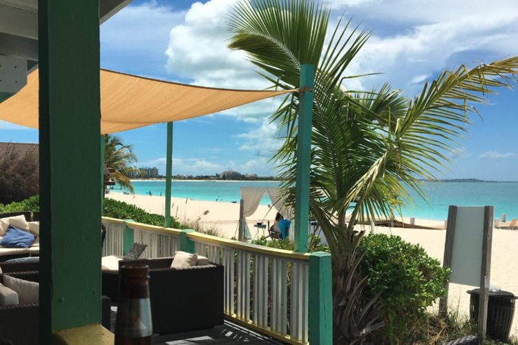 Beachside outdoor seating area with black wicker chairs and cushions, green palm tree, turquoise ocean, sandy beach, and blue sky with clouds.