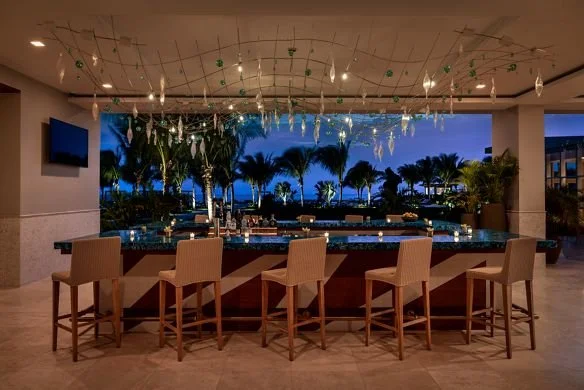 Indoor bar area with five beige chairs, decorated ceiling, and a view of palm trees and sky outside.