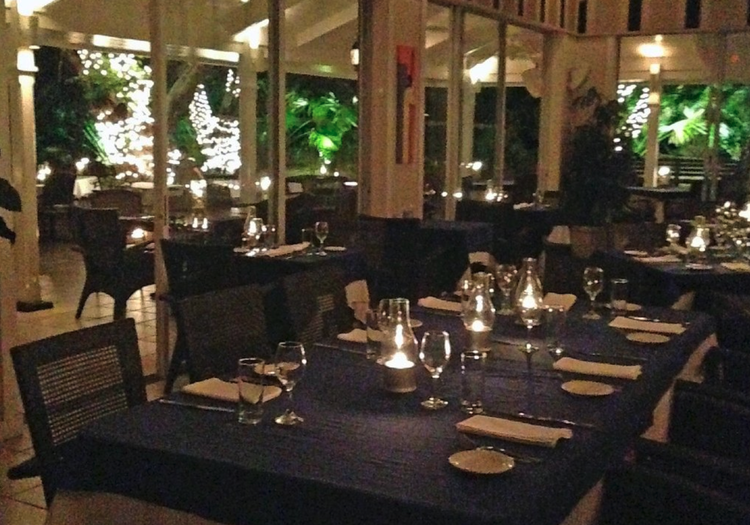 Dining area at a restaurant with tables covered in dark tablecloths, set with glasses, napkins, and plates, illuminated by candlelight, with large windows revealing greenery and Christmas lights outside.