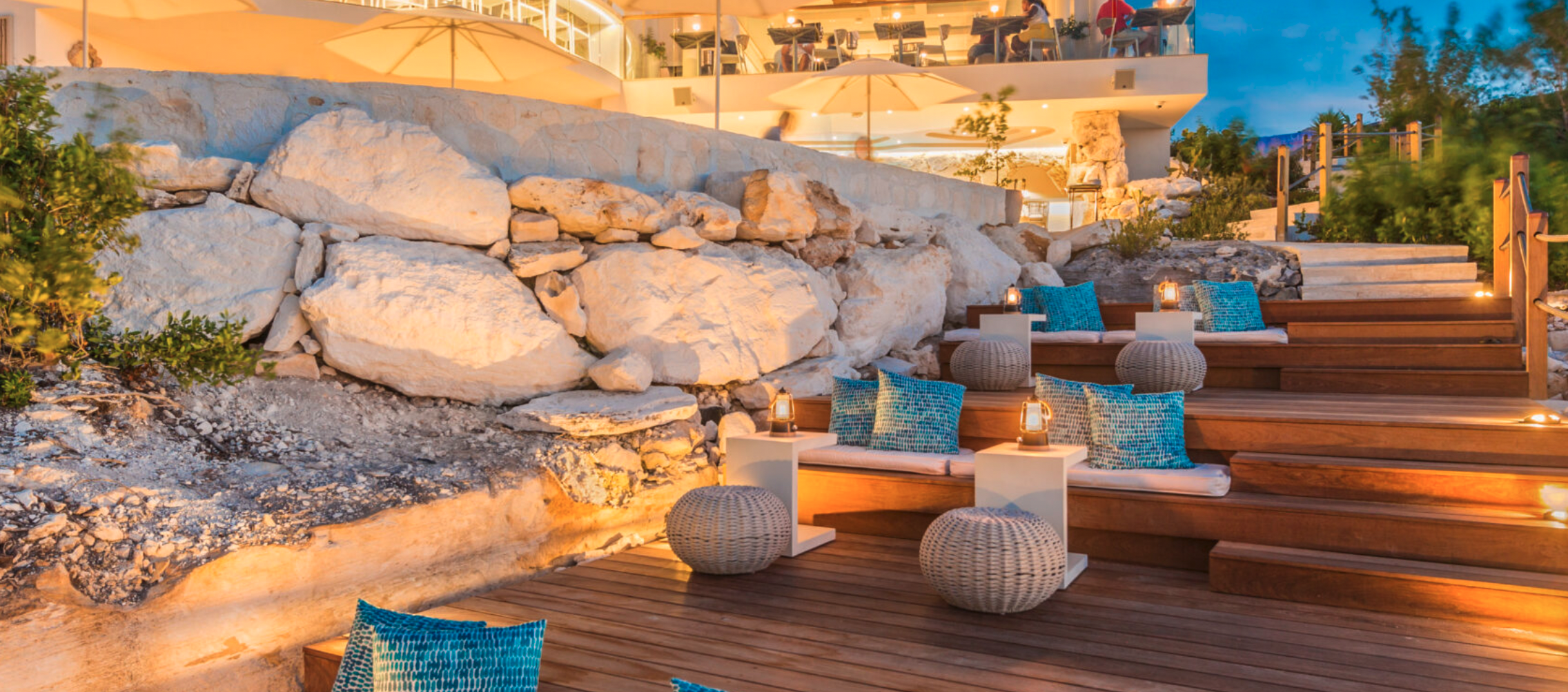 Outdoor seating area with wood benches, blue patterned cushions, round wicker stools with backrests, small white side tables, and lanterns, set on a wooden deck at dusk, with large white rocks and a modern building with glass balconies in the background.
