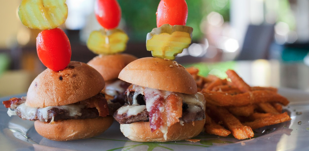 Mini sliders with cherry tomatoes, pickles, bacon, and cheese, served with sweet potato fries on a plate.