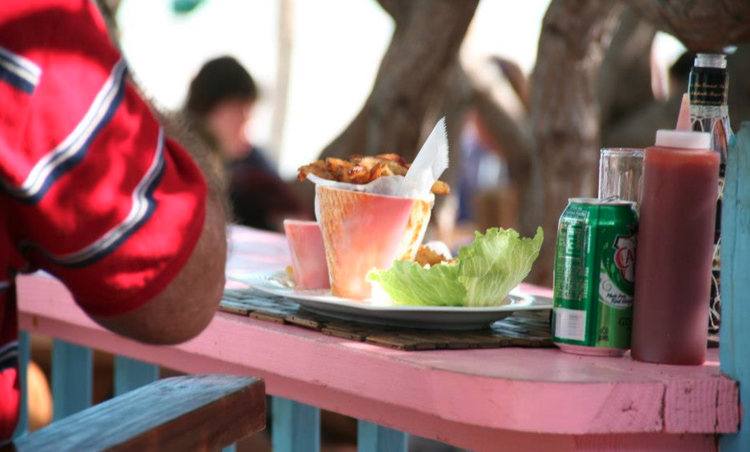 Close-up of a pink outdoor table with a plate of fried chicken, lettuce, and a drink, surrounded by drinks and condiments at a casual outdoor dining setting.