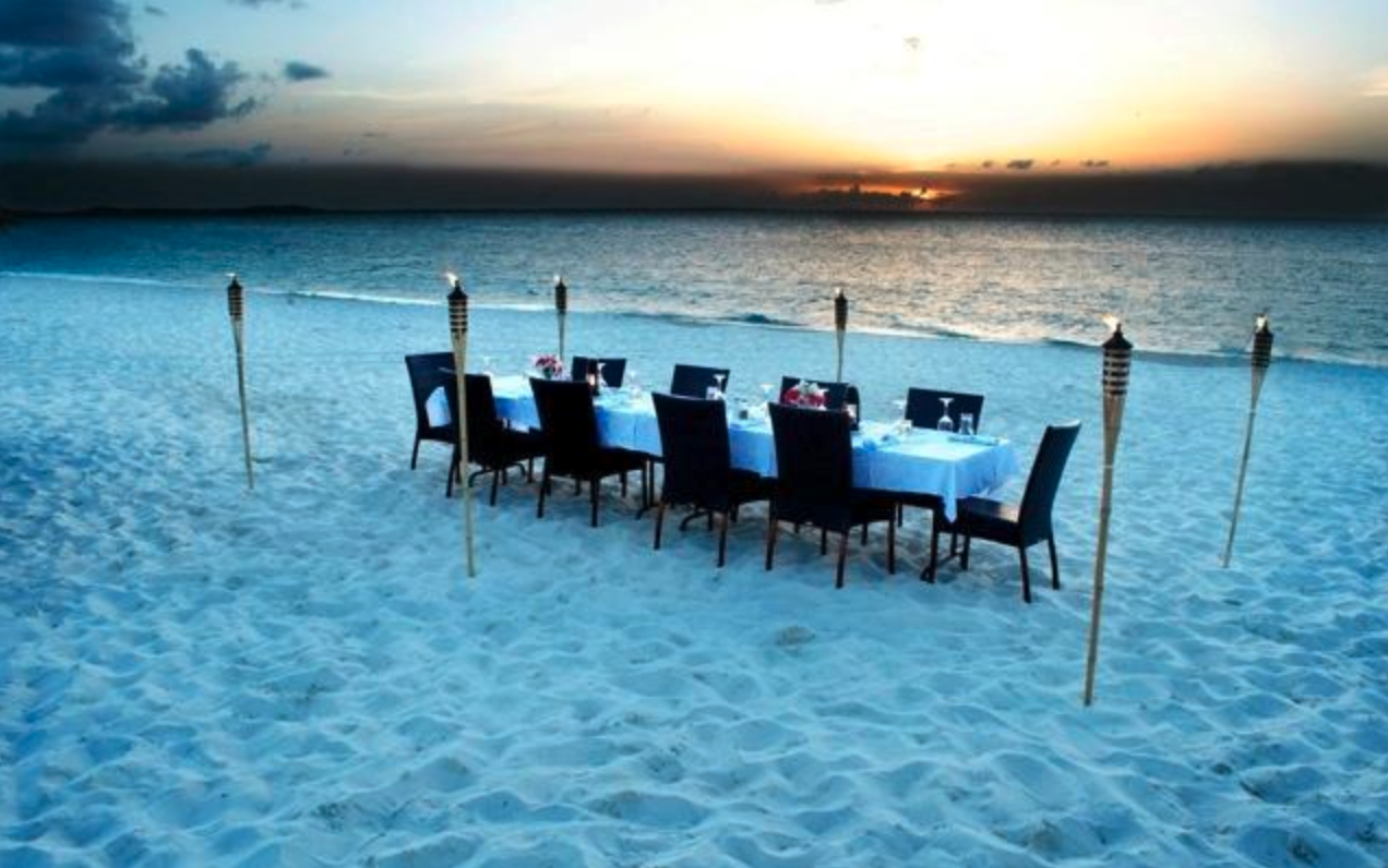 A dining table set for dinner on a beach with tiki torches around it, overlooking the ocean at sunset.