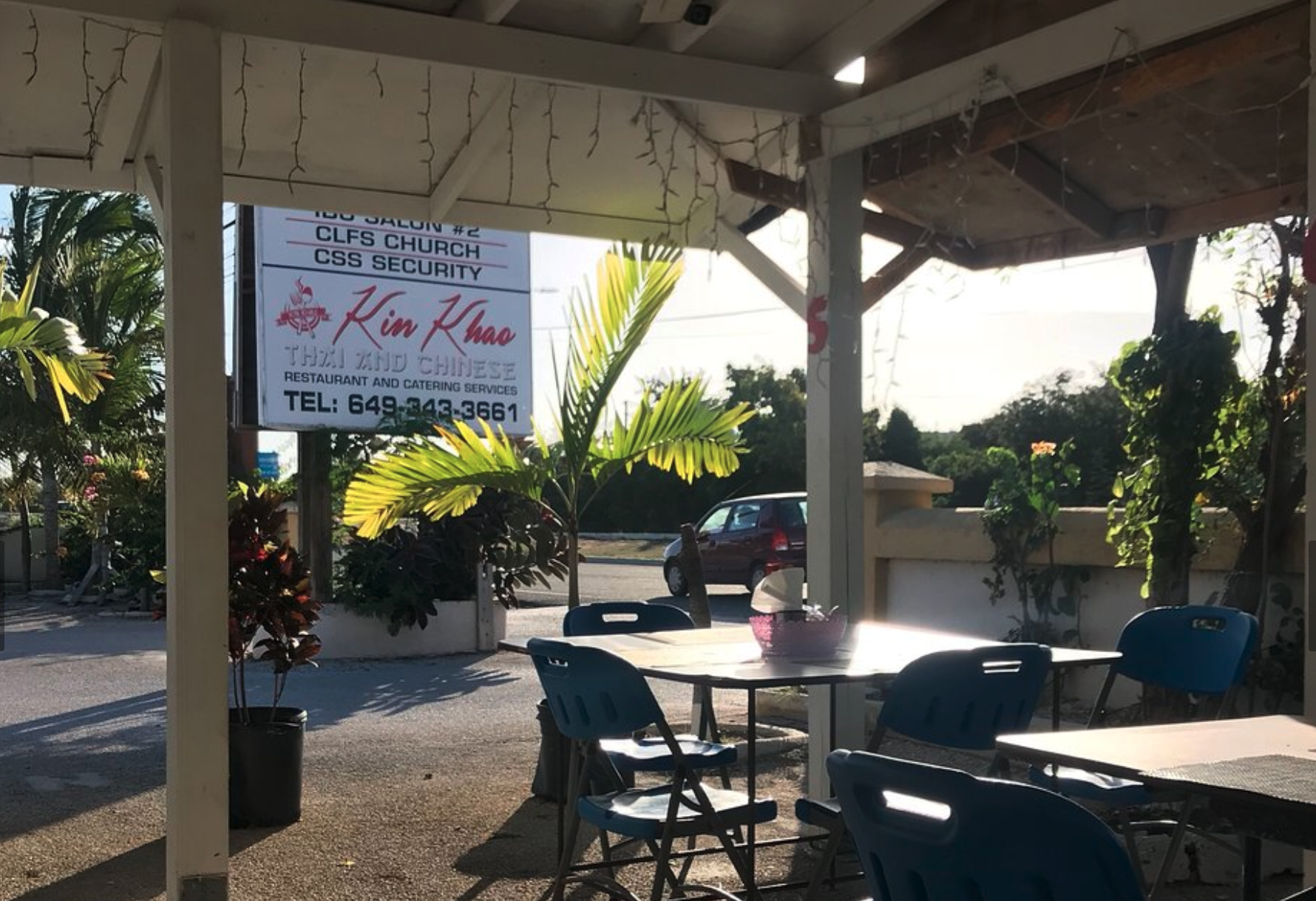 Outdoor seating area of a restaurant with plastic tables and chairs, potted plants, and a hanging sign displaying the restaurant's name 'Kim Khao' and contact information, with a view of parked cars and trees in the background.