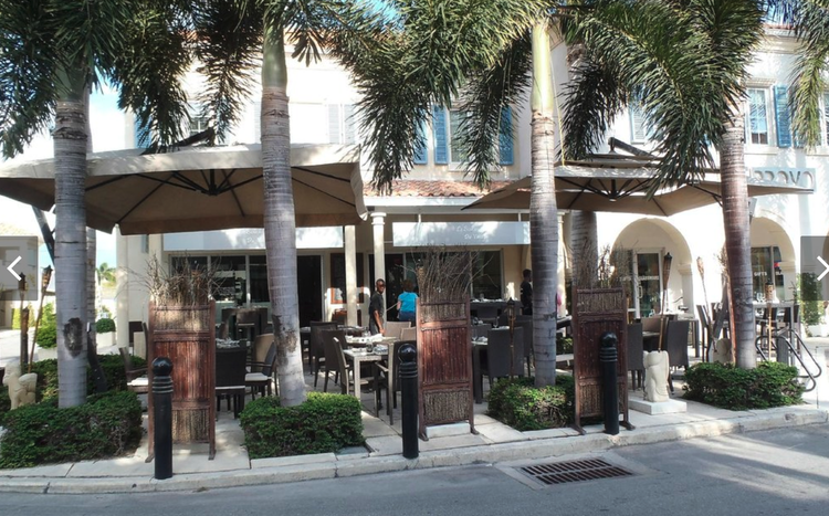 Outdoor restaurant seating area with tables, chairs, umbrellas, palm trees, and a building in the background.
