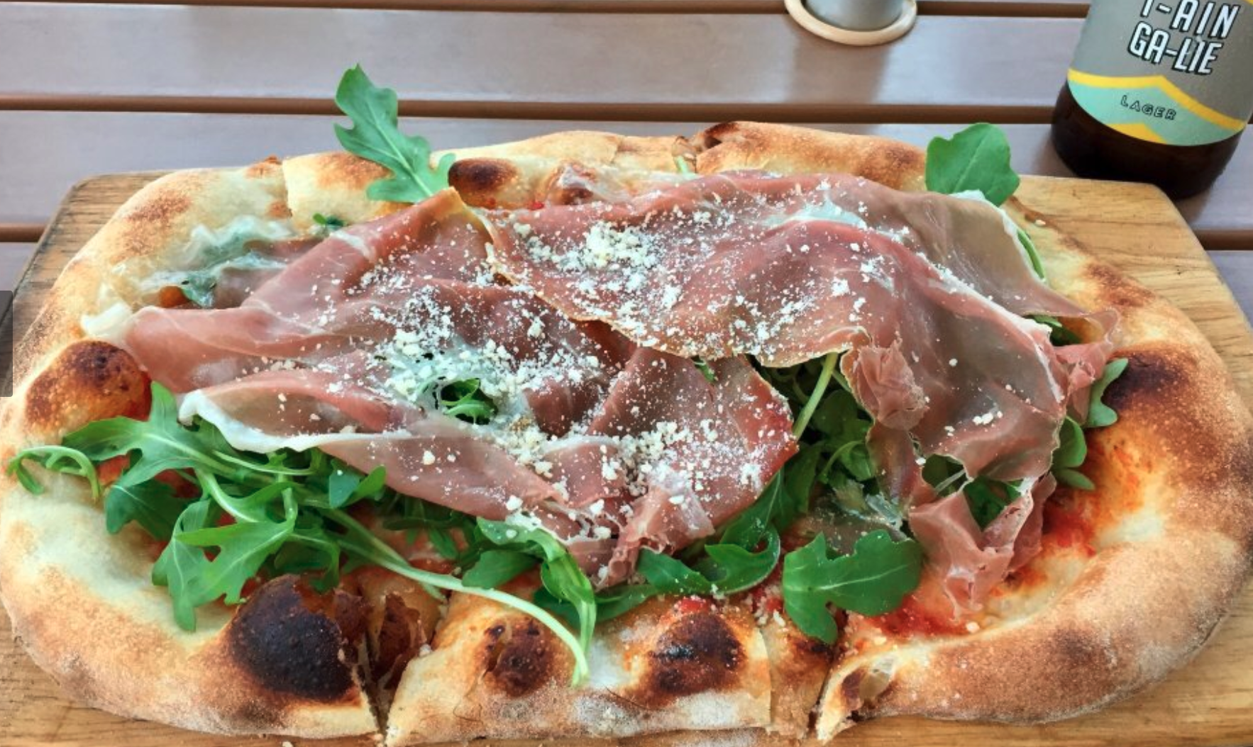 Pizza topped with arugula, prosciutto, and grated cheese on a wooden board, with a beer bottle in the background.