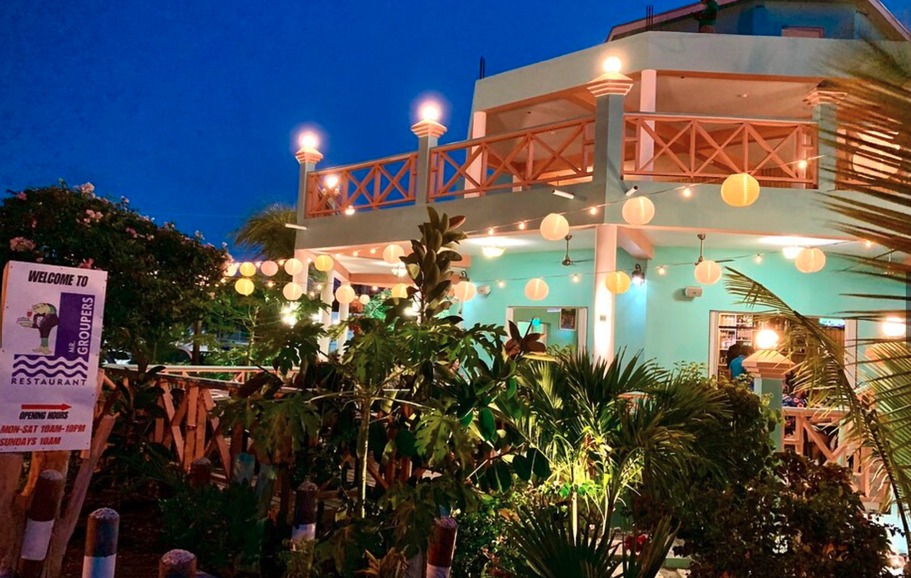 A two-story restaurant building decorated with hanging lanterns and string lights during dusk, surrounded by lush tropical plants and a welcome sign in the foreground.