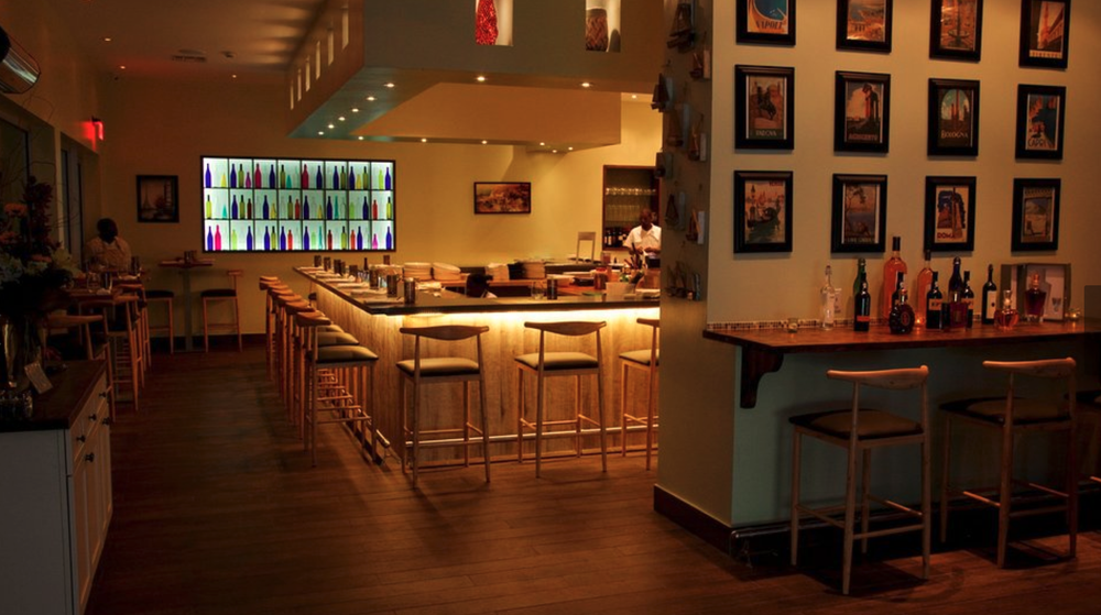 Interior of a modern bar with a U-shaped counter, bar stools, colorful bottle art display, and framed artwork on walls.
