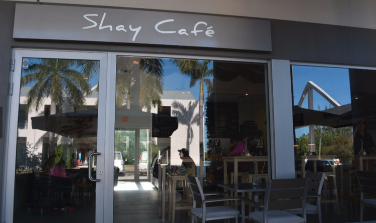 The exterior of Shay Café with glass doors and windows, showing palm trees and outdoor seating with tables and chairs, and a few people inside.
