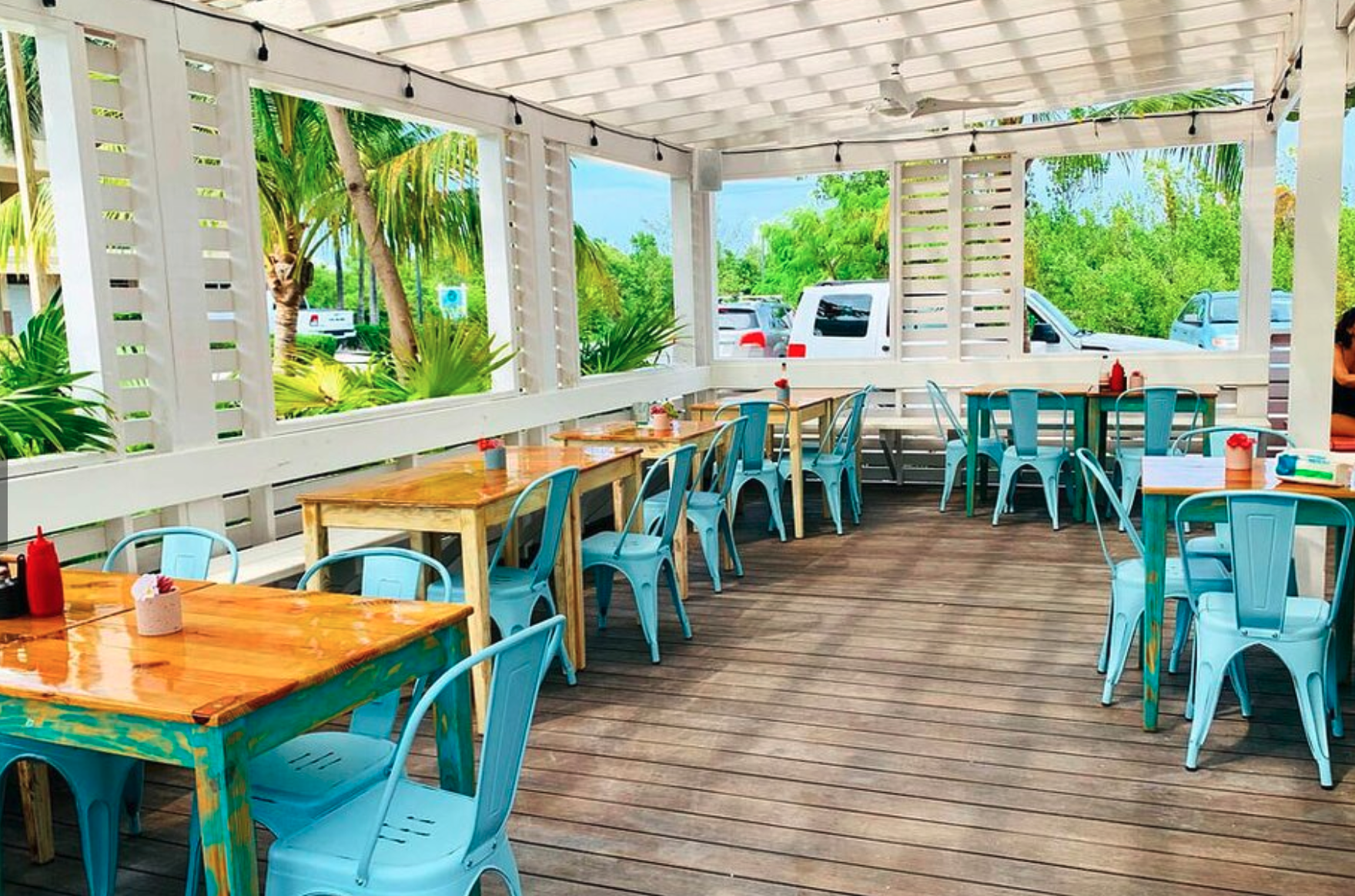 Empty outdoor restaurant patio with wooden tables, blue chairs, and beach greenery in the background.