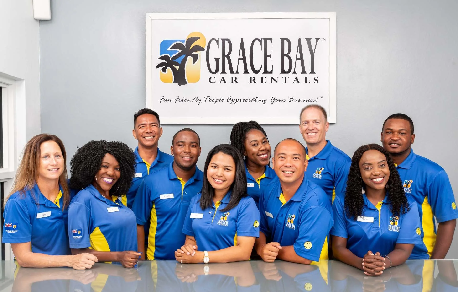 Group of employees wearing blue and yellow Grace Bay Car Rentals uniforms posing inside an office with a sign that reads 'Grace Bay Car Rentals' and the slogan 'Fun Friendly People Appreciating Your Business'.