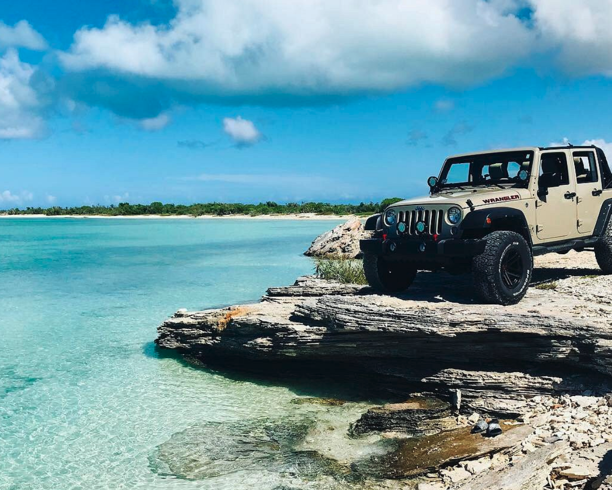 A beige Jeep Wrangler parked on rocky shoreline by clear blue water with green vegetation and partly cloudy sky in the background.