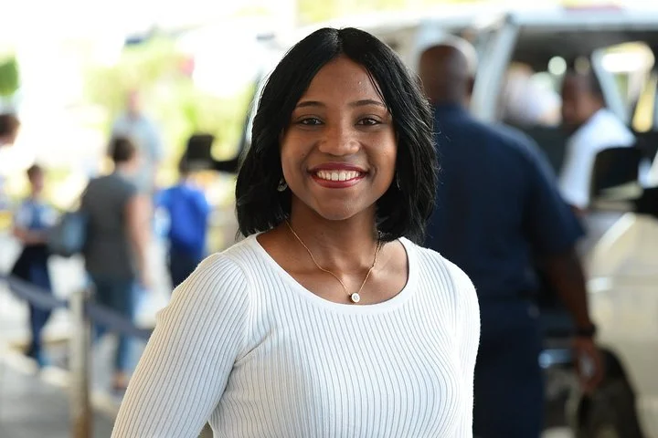 A young woman with shoulder-length black hair, wearing a white ribbed long-sleeve shirt and a necklace, smiling outdoors in a busy area with people and vehicles in the background.