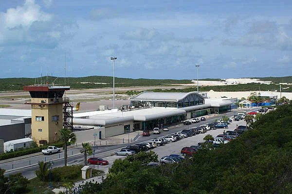 View of an airport terminal with a control tower and parked cars, situated next to sandy dunes and a cloudy sky.
