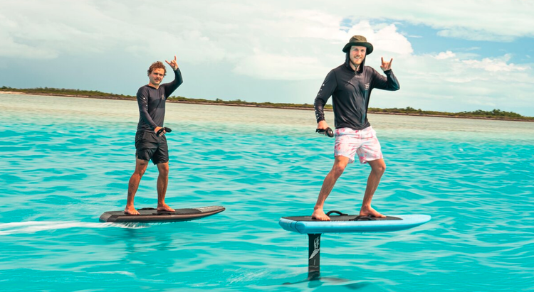 Two men stand on paddleboards in turquoise water, making peace signs, with a sandy shoreline and partly cloudy sky in the background.