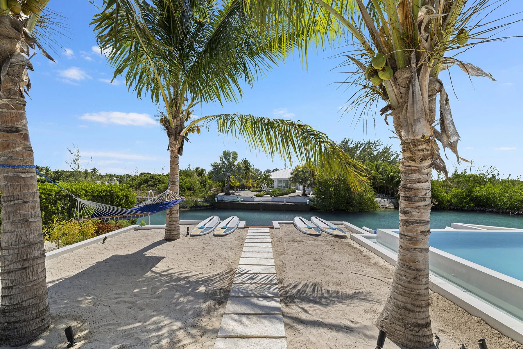 Beachside scene with two palm trees, a hammock between them, three paddleboards on sand, a stepping stone path, clear blue water, and a house in the background.